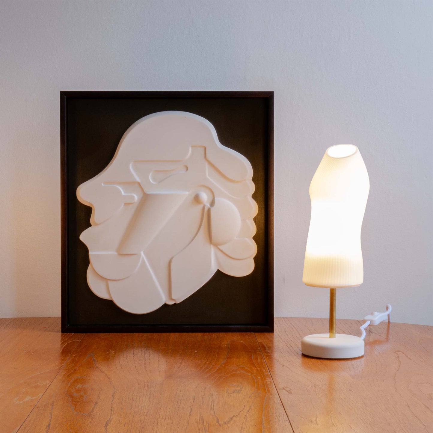 The Figuratives bundle includes the Ora lamp with a brass rod and Mask 1 framed in Mahogany stain. The lamp and relief are photographed on a wooden table