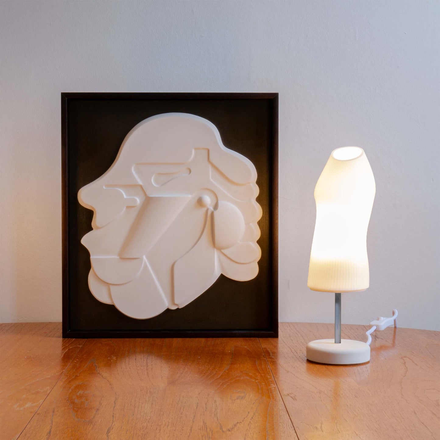 The Figuratives bundle includes the Ora lamp with a chrome rod and Mask 1 framed in Mahogany stain. The lamp and relief are photographed on a wooden table.