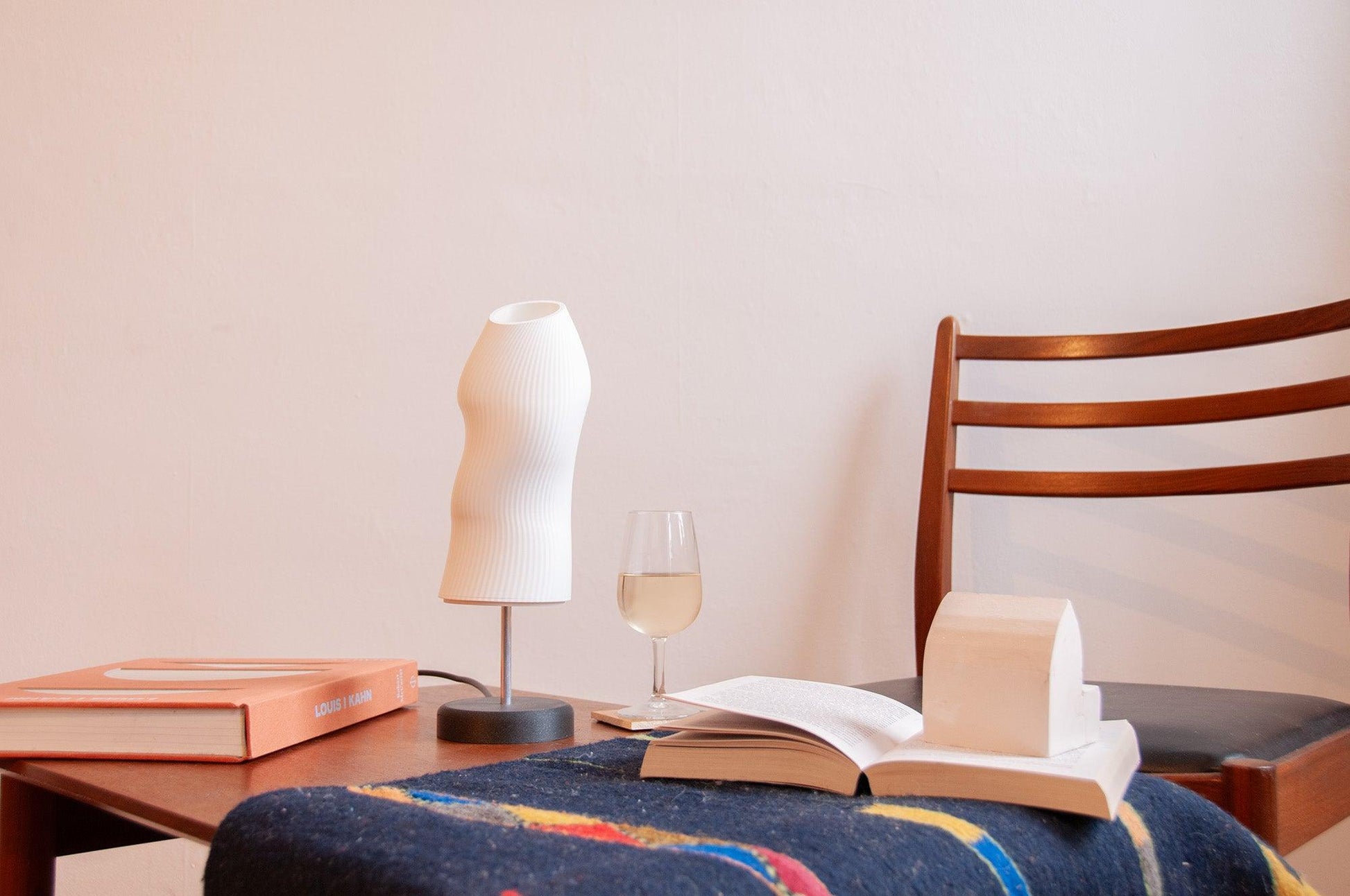 Ora sculptural table lamp positioned in a living room on a wooden table next to a glass of white wine and a book